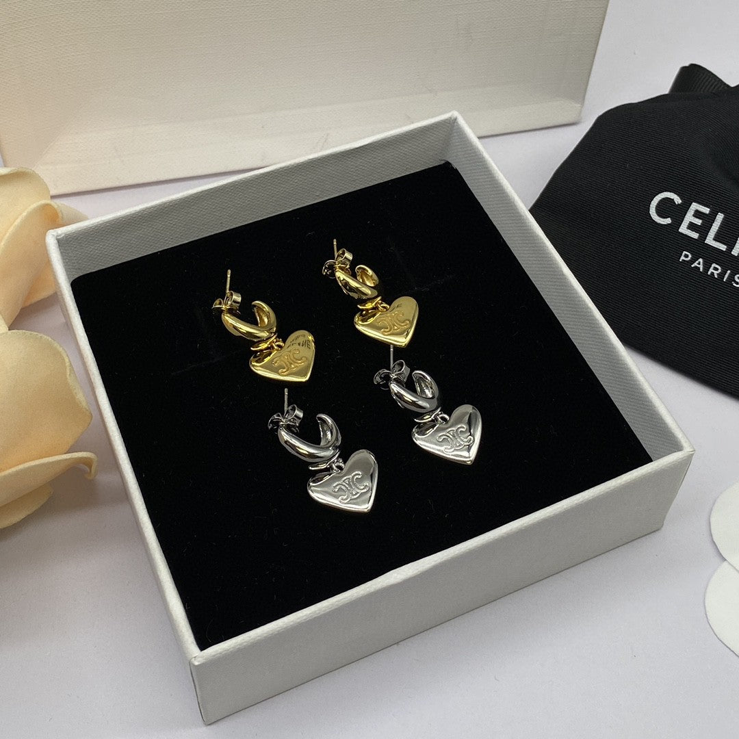 A pair of heart-shaped dangle earrings displayed in an open jewelry box. One earring is gold-toned and the other is silver-toned, both featuring a curved top element and a dangling heart charm engraved with the word 'LOVE'.