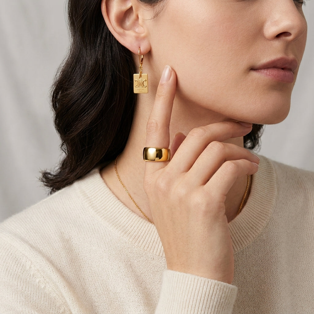 A close-up lifestyle shot of a model wearing a gold square drop earring with a small engraved monogram and a wide gold band ring, styled with a cream knit sweater.
