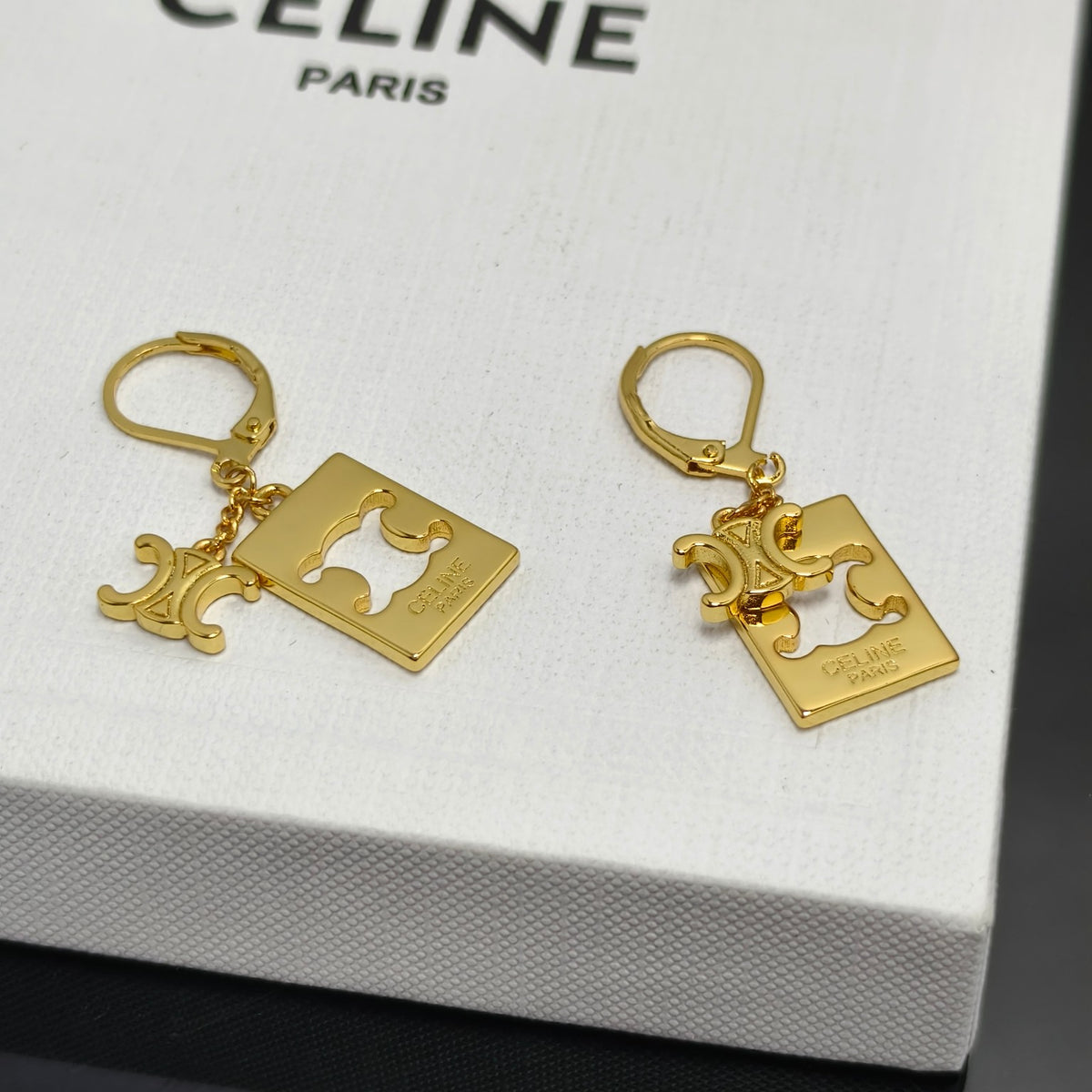 A pair of gold-tone Celine leverback earrings with a square logo charm and a small Celine Triomphe charm, displayed on a white Celine box.