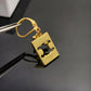 A gold-colored metal bottle opener keychain with a lobster clasp, held by metal tweezers against a dark reflective surface. The brand name 'CURE' and 'SWISS' are engraved on the side of the rectangular opener.