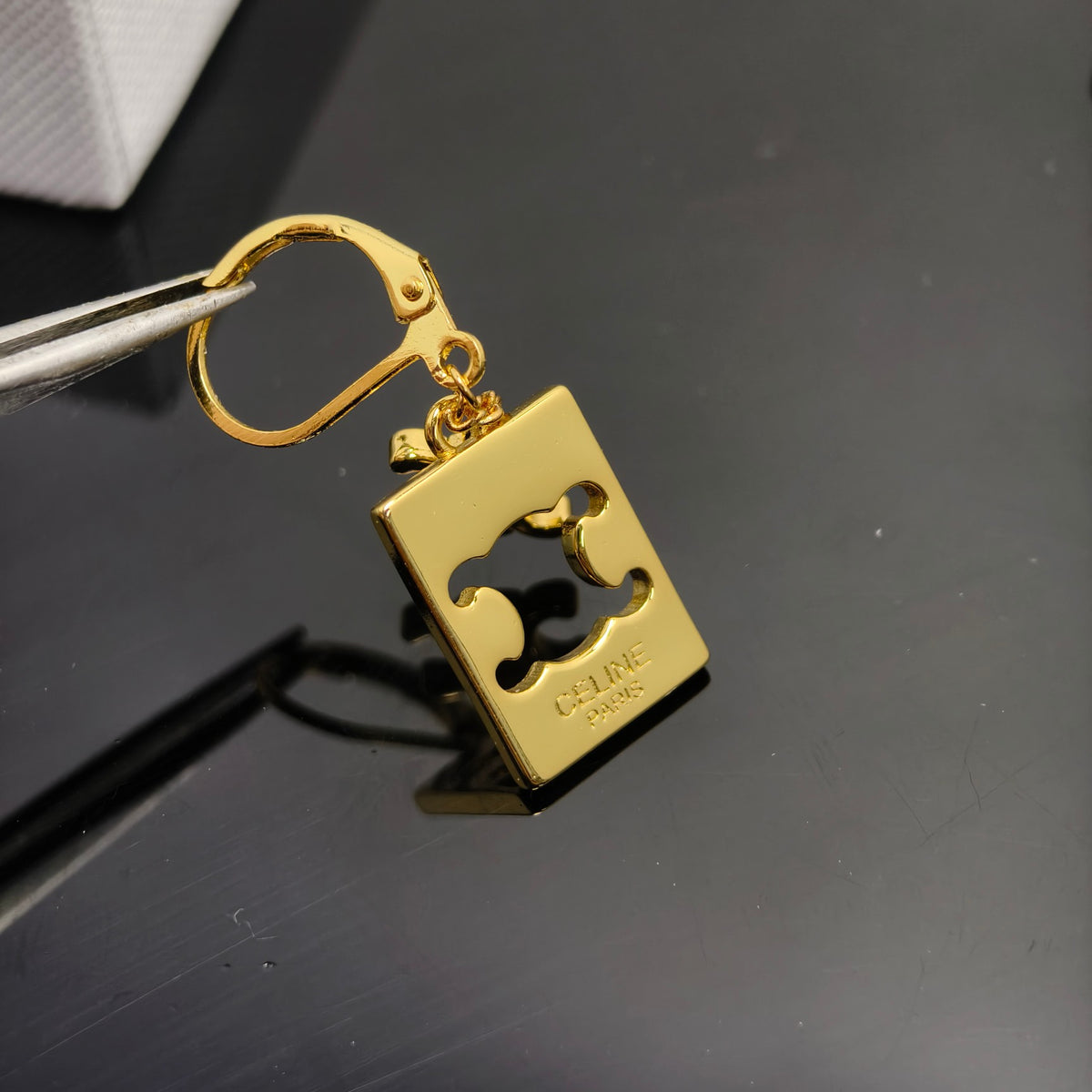 A gold-colored metal bottle opener keychain with a lobster clasp, held by metal tweezers against a dark reflective surface. The brand name 'CURE' and 'SWISS' are engraved on the side of the rectangular opener.