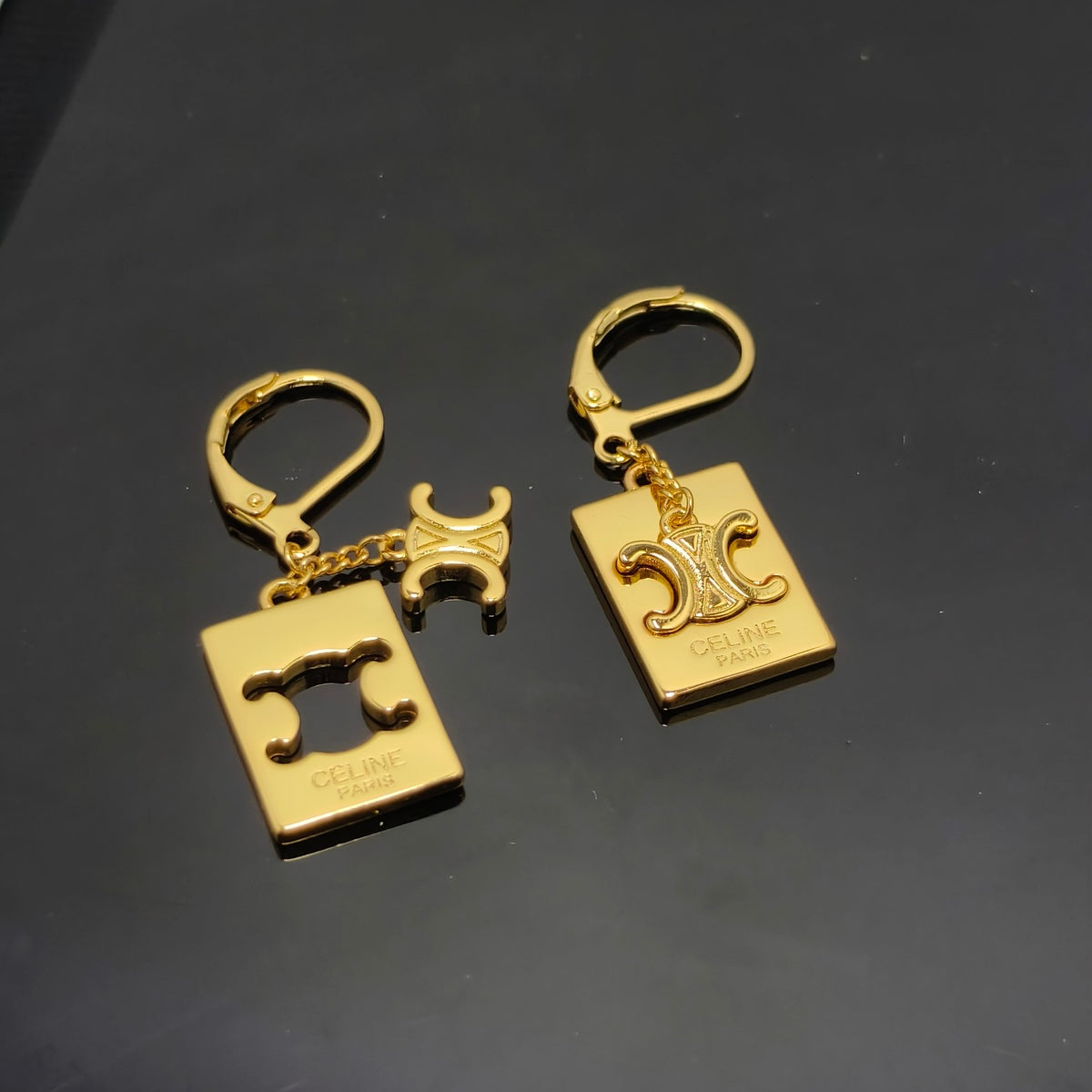 Two gold-tone Celine keychains displayed on a dark reflective surface. Each keychain features a square charm with the Celine Triomphe logo, a small horse-shaped charm, and a lobster clasp.