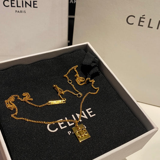 A gold-tone Céline necklace with a small rectangular pendant and matching charm lies on a black Céline box insert inside an open Céline box, with branded white packaging visible in the background.