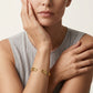 A close-up of a model wearing a gold bangle bracelet with a textured geometric pattern. The model is wearing a light gray pleated top and posing with one hand on her cheek against a clean, off-white background.