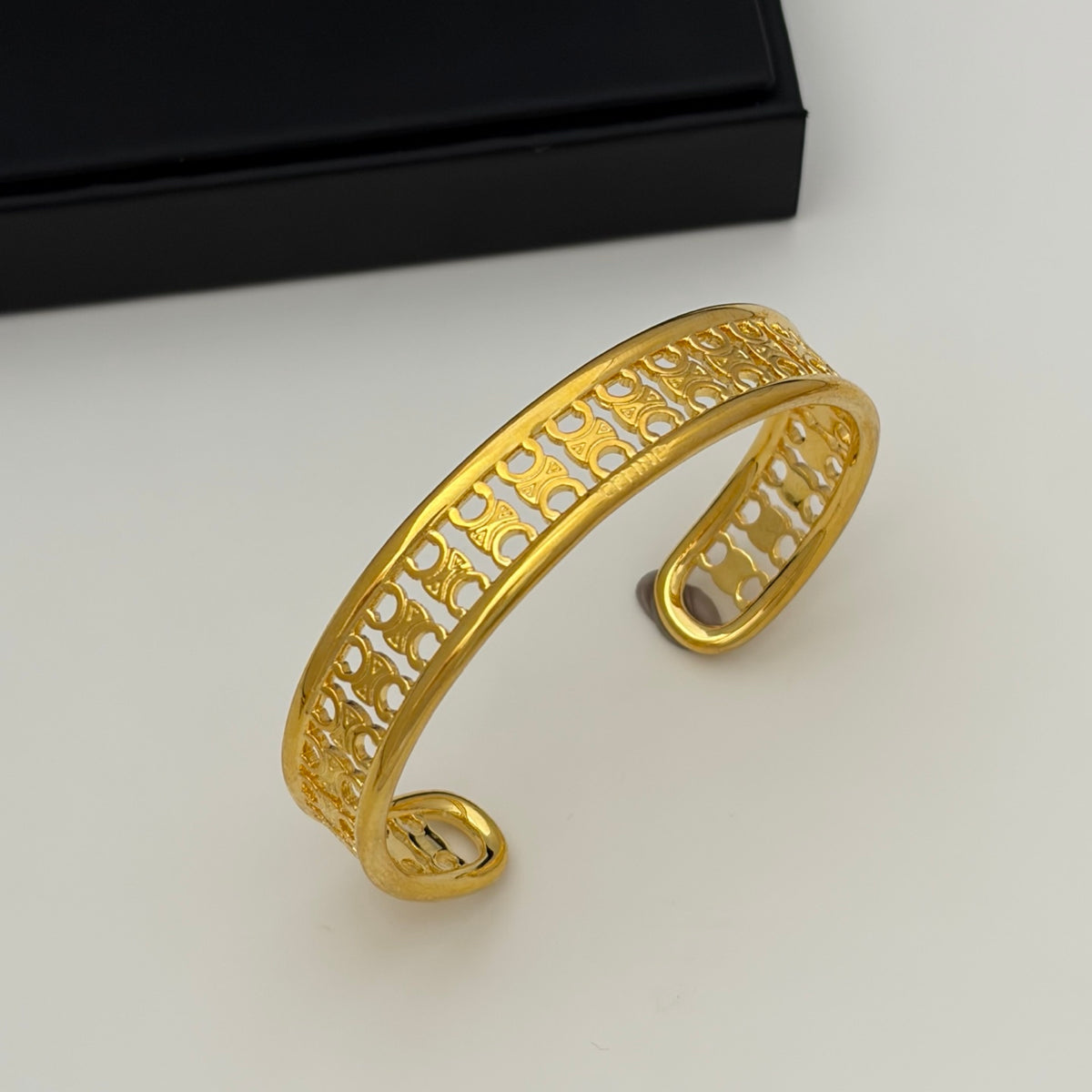 A gold-toned filigree cuff bracelet with an openwork pattern, displayed on a light gray surface with a black box in the background.