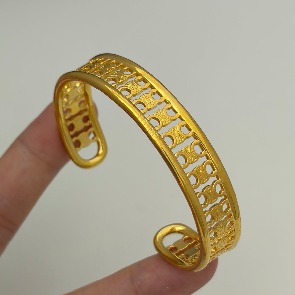 A gold-plated metal cuff bracelet with intricate openwork patterns, held by two fingers to show its scale against a plain light gray background.