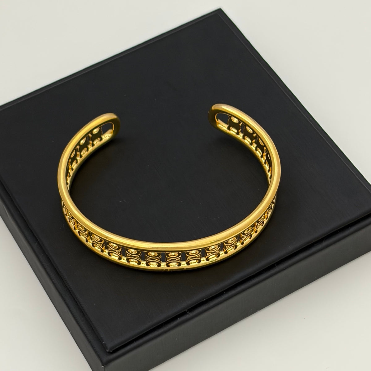 A gold-toned open cuff bracelet with a textured beaded border, displayed on a black presentation box against a light gray background.