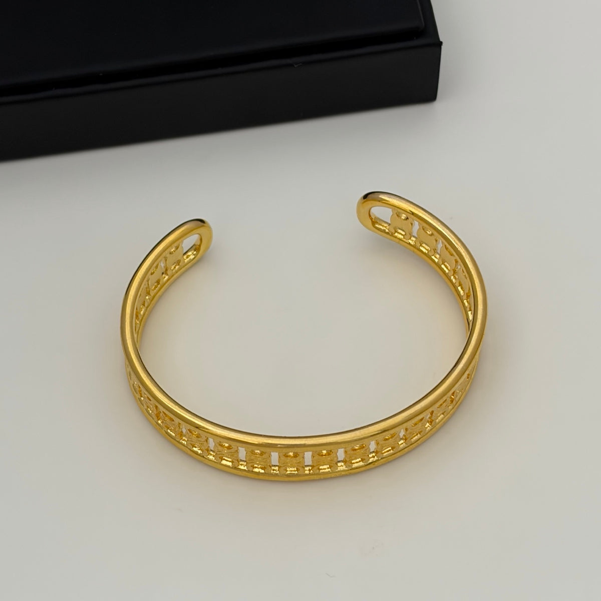 A gold-plated open cuff bracelet with a detailed geometric pattern along the front, displayed on a light gray surface with a black box in the background.