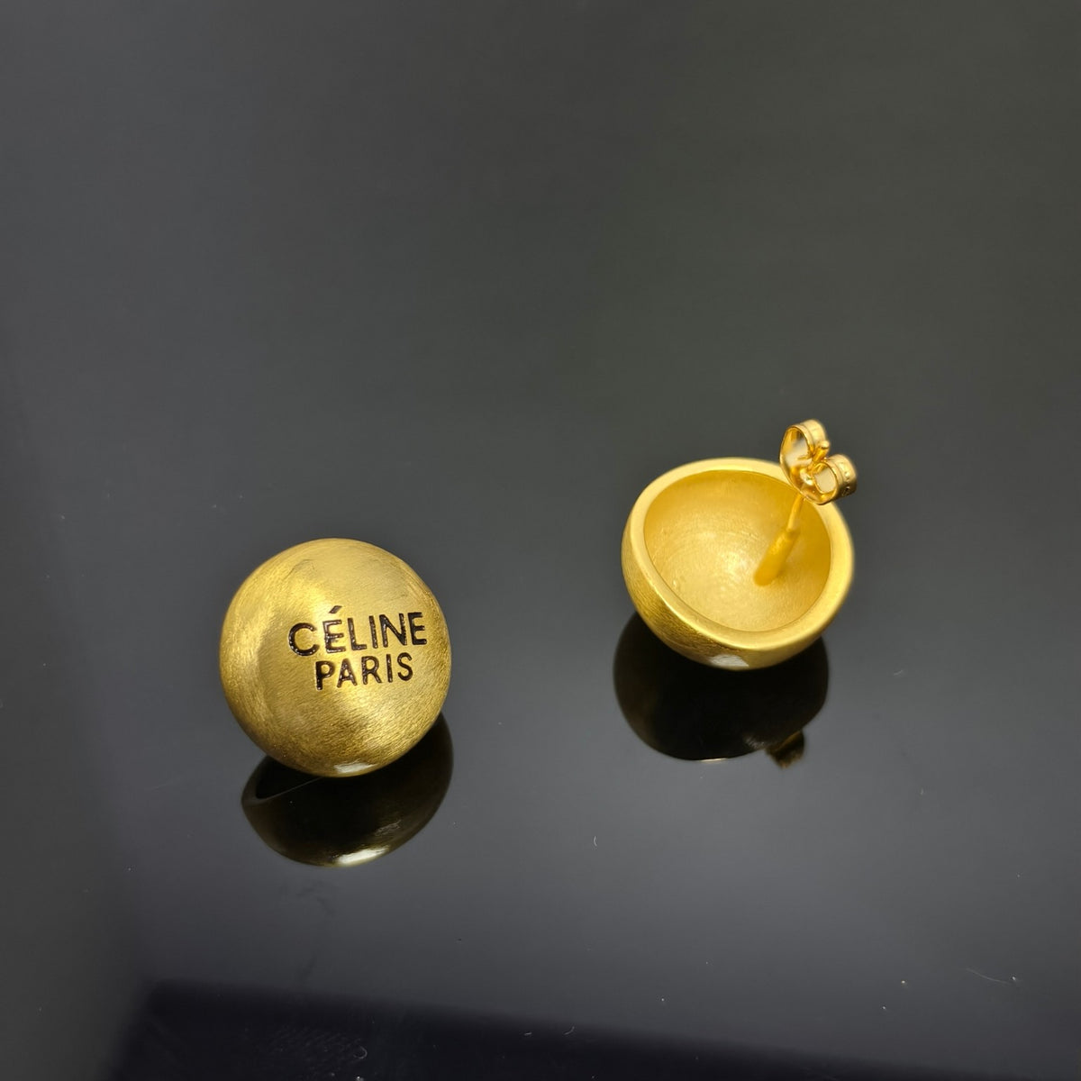 A pair of gold-tone Celine Paris round stud earrings shown on a reflective black surface, with one earring facing forward displaying the engraved brand name and the other showing the back with a post and clasp.
