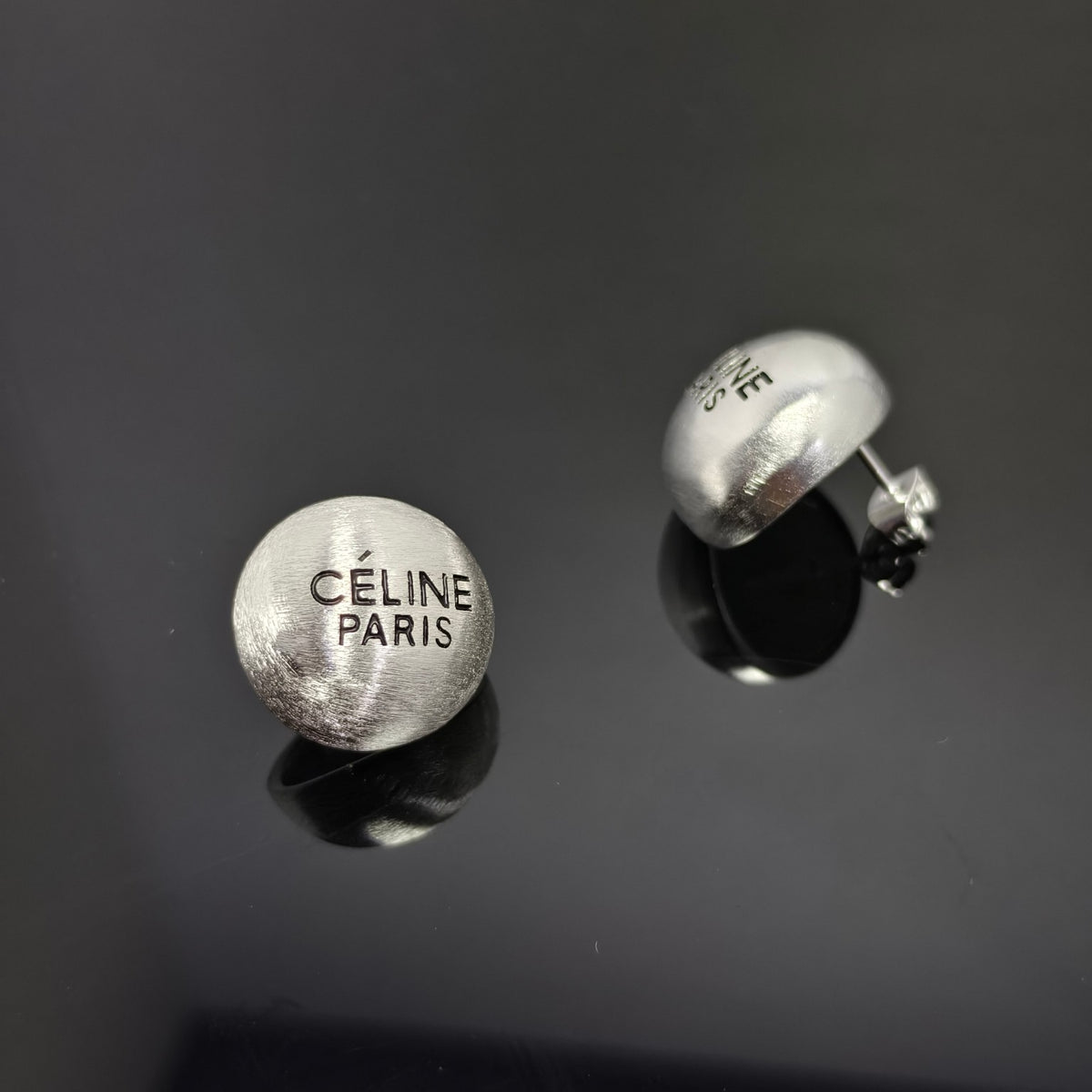 A pair of silver-tone oval stud earrings engraved with 'CÉLINE PARIS', displayed on a dark reflective surface.