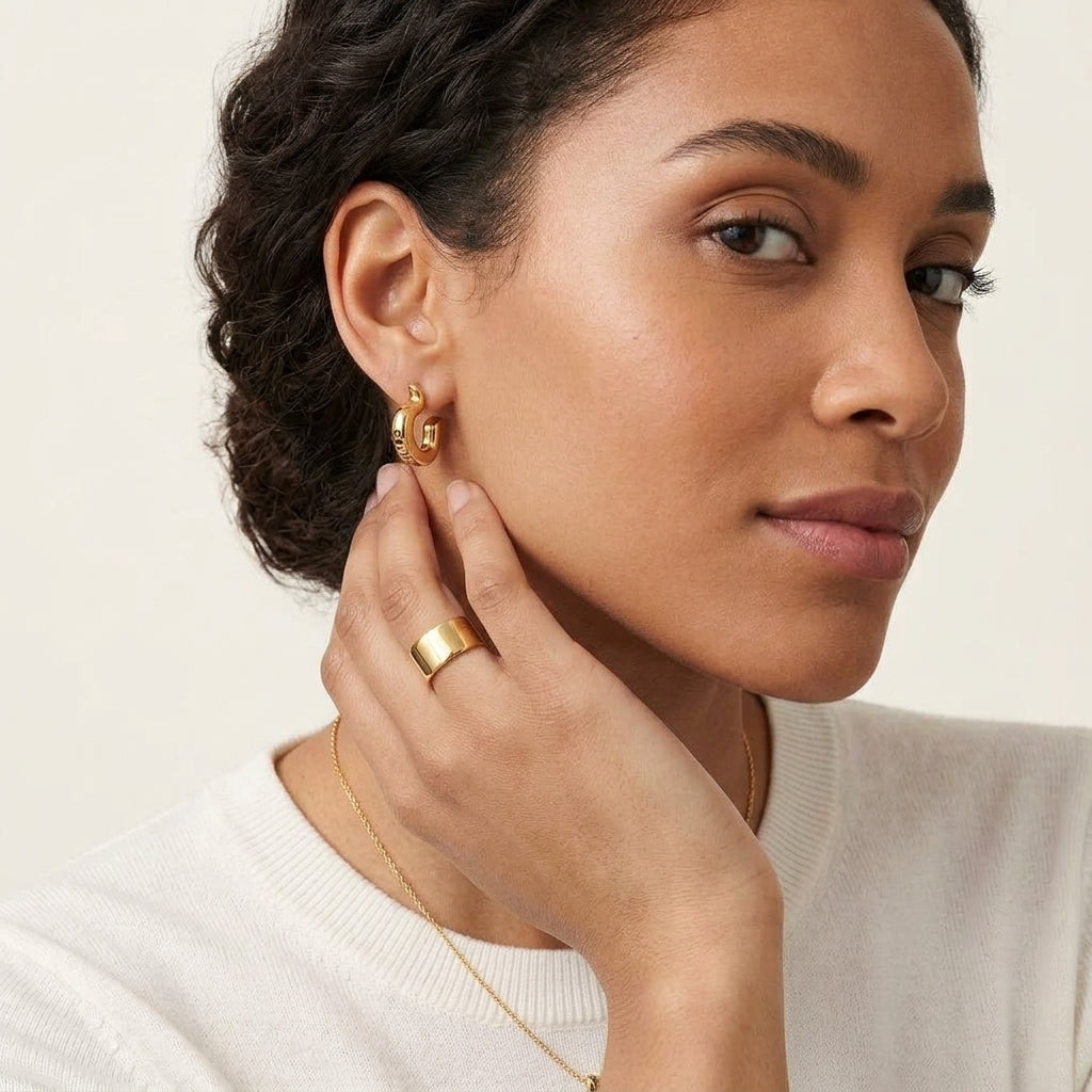 A close-up lifestyle shot of a woman wearing a delicate gold necklace with a circular interlocking logo pendant, along with a matching gold hoop earring and a gold ring, against a neutral beige background.