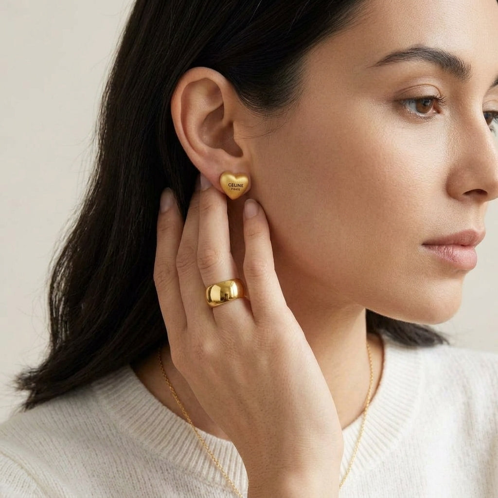 A close-up of a woman wearing a small gold heart stud earring and a matching gold ring, with a delicate gold necklace visible against a soft light background.
