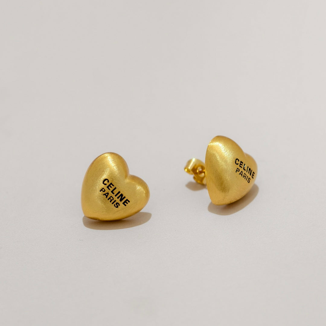 A pair of gold heart-shaped stud earrings engraved with 'CELINE PARIS' in black lettering, displayed on a clean light gray background.