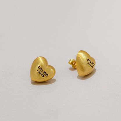 A pair of gold heart-shaped stud earrings engraved with 'CELINE PARIS' in black lettering, displayed on a clean light gray background.