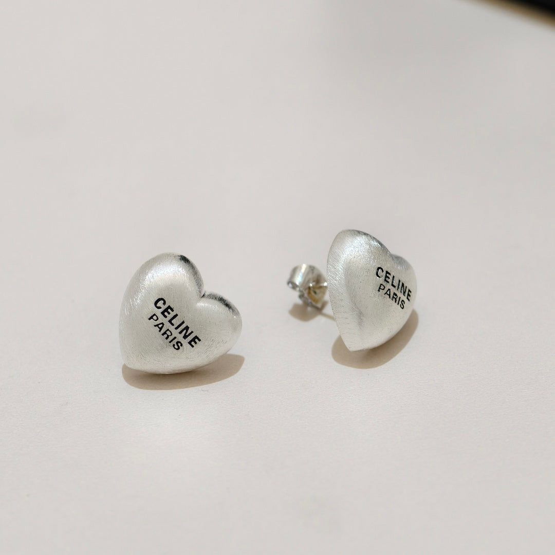 A pair of silver heart-shaped stud earrings engraved with 'CELINE PARIS', displayed on a clean, light beige surface.
