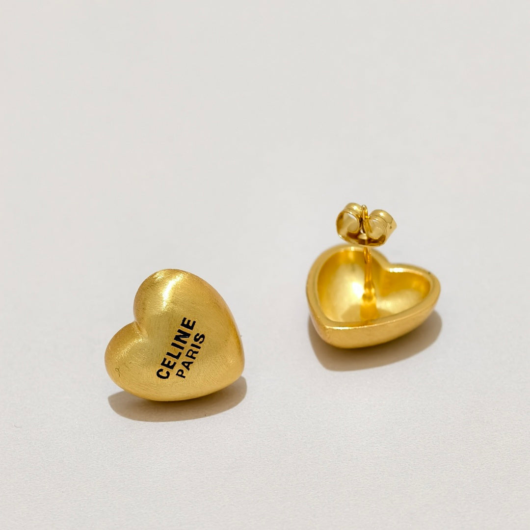 Two gold heart-shaped stud earrings on a light gray background, one showing the front with 'CELINE PARIS' printed in black and the other showing the back with a butterfly clasp.