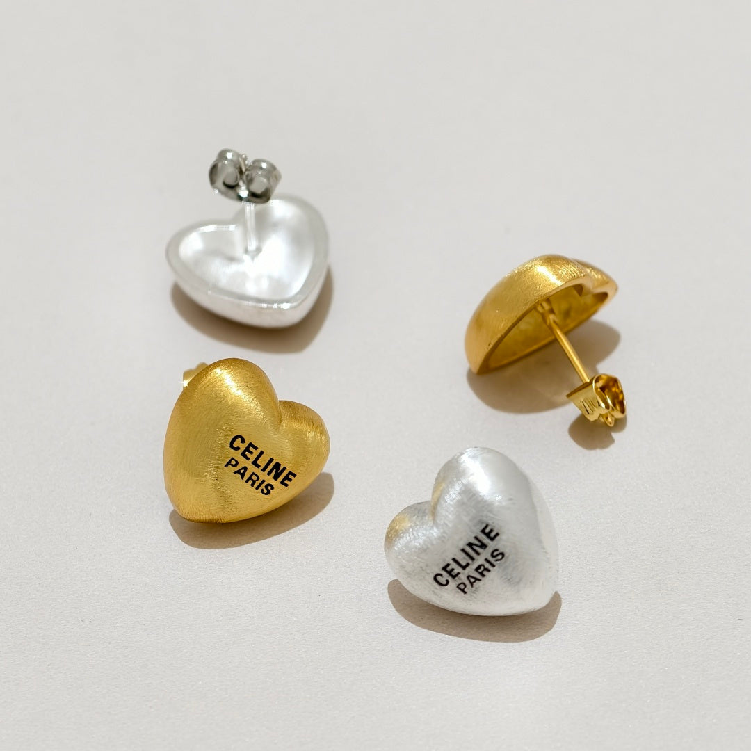 Four heart-shaped Celine stud earrings in polished gold and silver, showing both front and side/back views on a light beige background.