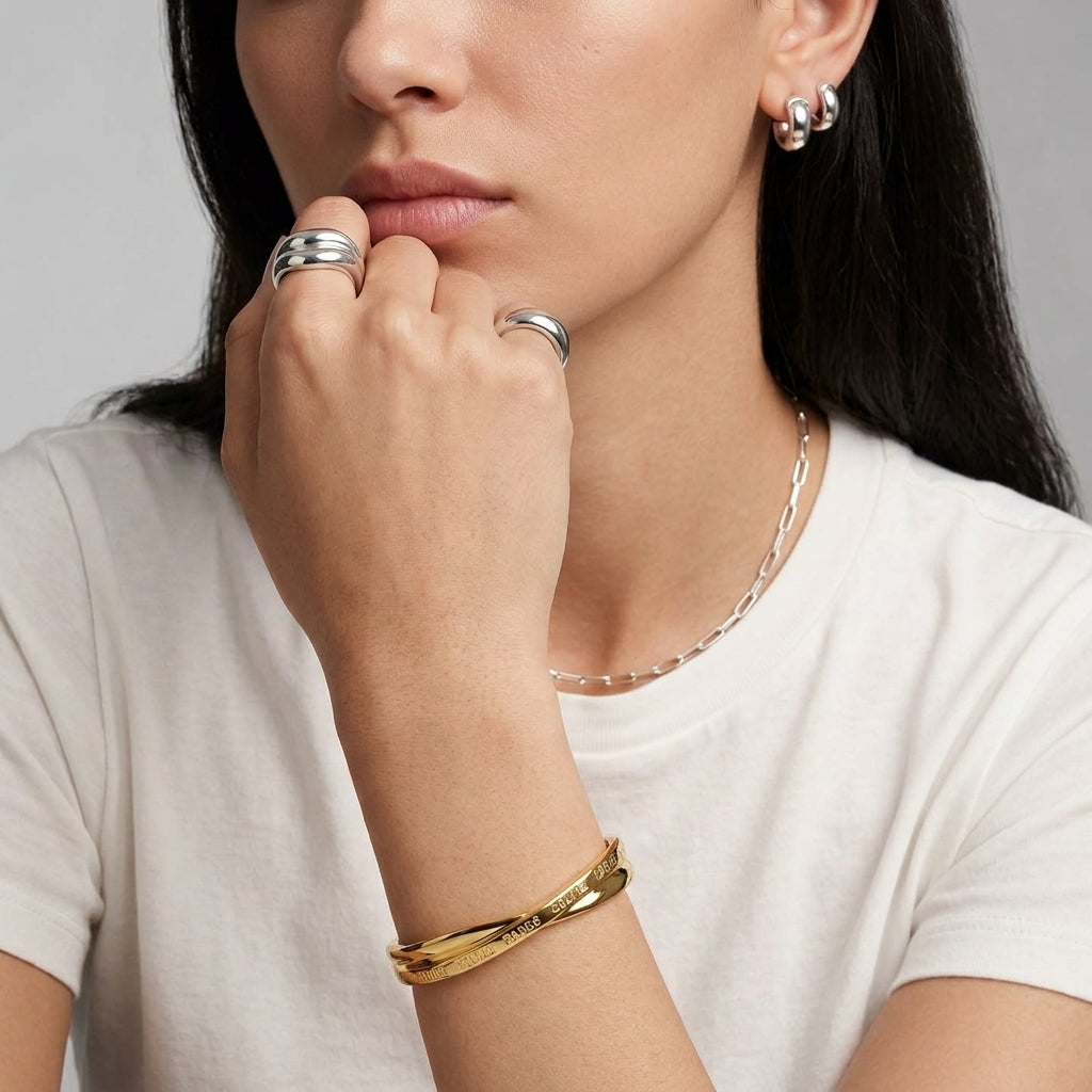 A close-up lifestyle shot of a model wearing a gold twisted bangle bracelet, silver rings, and a silver chain necklace, paired with a white t-shirt against a neutral gray background.