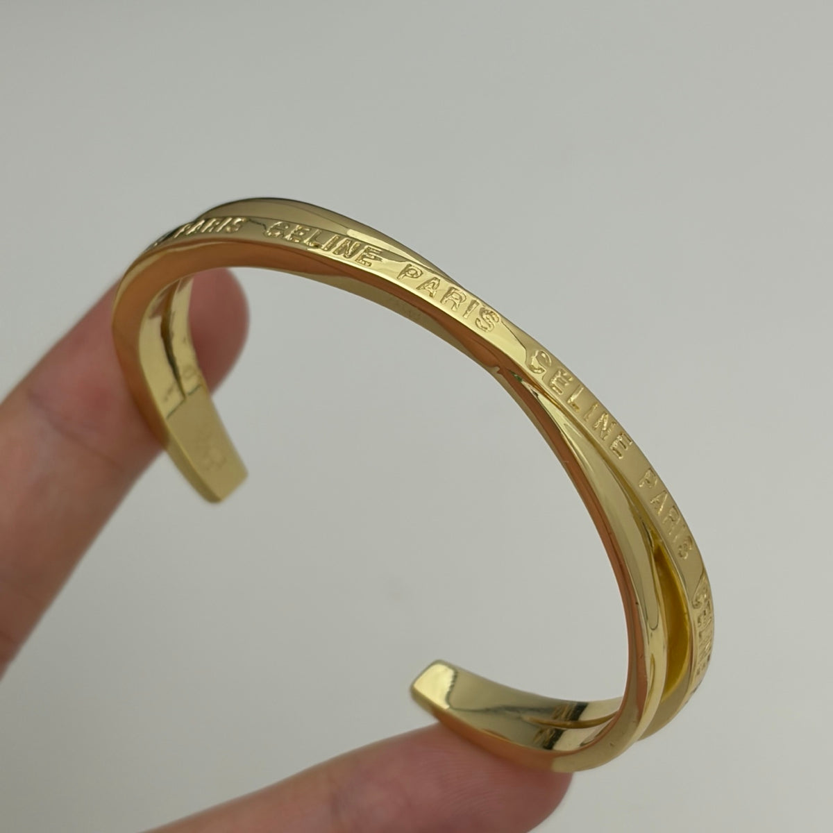 A gold-tone metal cuff bracelet with engraved 'CELINE PARIS' text, held by a person's fingers against a plain light gray background.