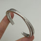 A silver-toned Celine cuff bracelet held by two fingers against a plain light gray background. The polished metal is engraved with 'CELINE PARIS' repeatedly.