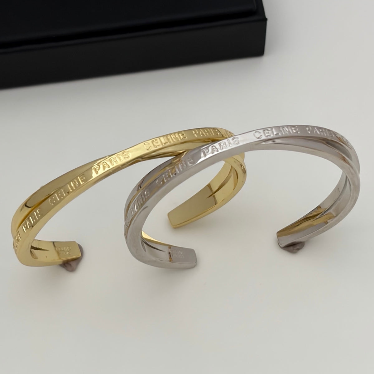 Two open cuff bracelets, one gold and one silver, engraved with 'CELINE PARIS', displayed on a light gray surface with a black box in the background.
