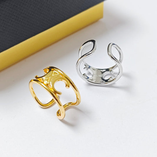 Two open cuff rings with a twisted abstract design, one in polished yellow gold and one in polished silver, displayed on a white surface with a yellow and black textured block in the background.