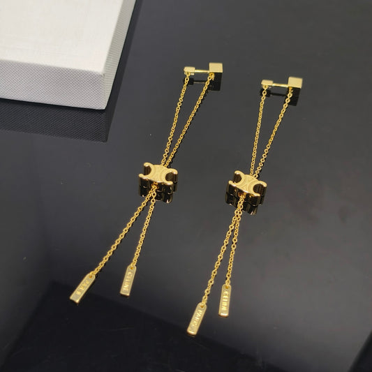 A pair of gold-tone dangle earrings displayed on a dark reflective surface, with a white box partially visible in the upper left corner.
