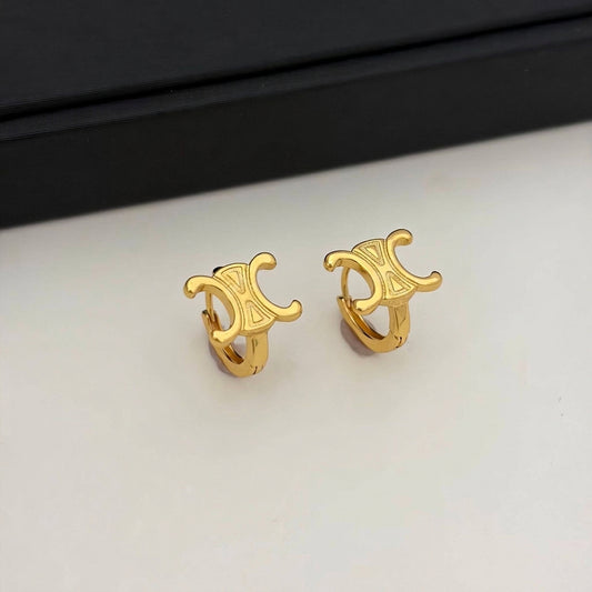 A pair of gold huggie hoop earrings with a stylized interlocking double-C logo design, resting on a white surface with a black box in the background.