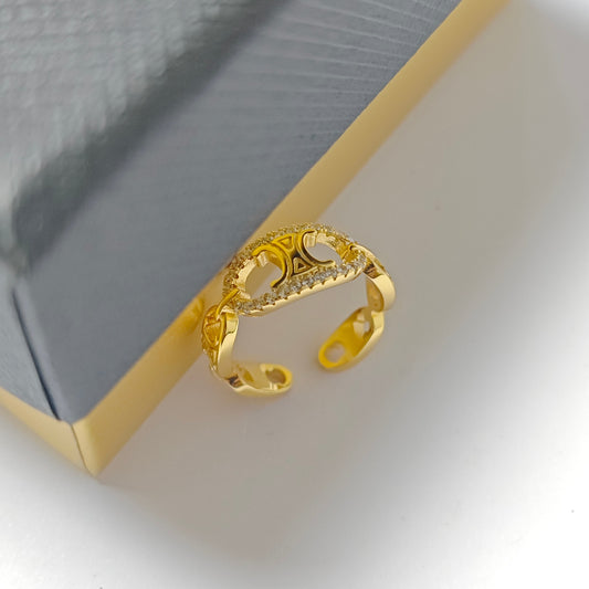 A gold-plated open ring with a decorative heart-shaped design and small clear stones, resting on a gray and gold textured jewelry box.