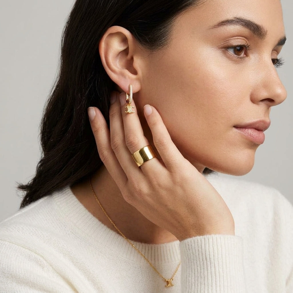 A close-up lifestyle shot of a woman wearing gold jewelry, featuring a small gold hoop earring with a starburst charm, a wide gold band ring, and a delicate gold necklace with a small star pendant, set against a soft gray background.