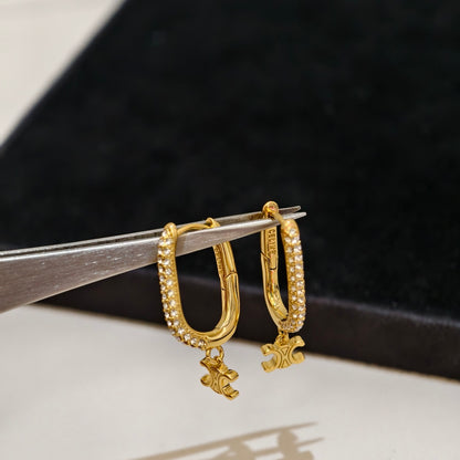 A pair of gold hoop earrings with pavé-set clear stones and small dangling charms, held on metal tweezers against a dark velvet jewelry display.