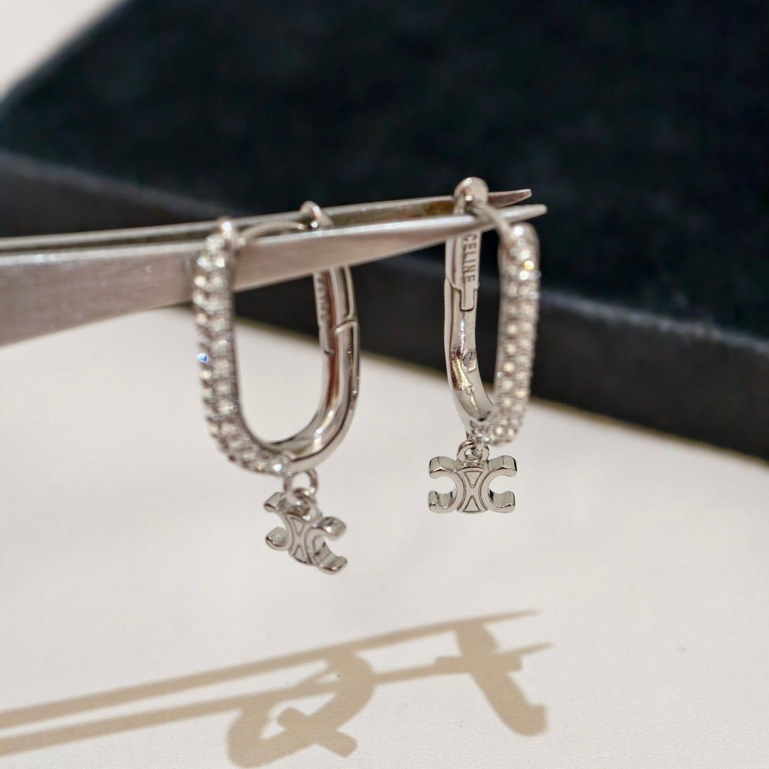 A pair of silver hoop earrings with small clear stones and dangling monogram charms, held by metal tweezers against a dark velvet surface and light background.