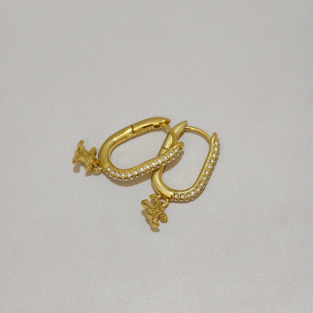 A pair of gold huggie hoop earrings adorned with small clear pavé stones and small dangling cherub charms, displayed on a neutral light gray background.