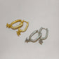 Two small hoop earrings on a plain light background, one in yellow gold and one in silver, each with a curved pavé-set band and a dangling animal charm.
