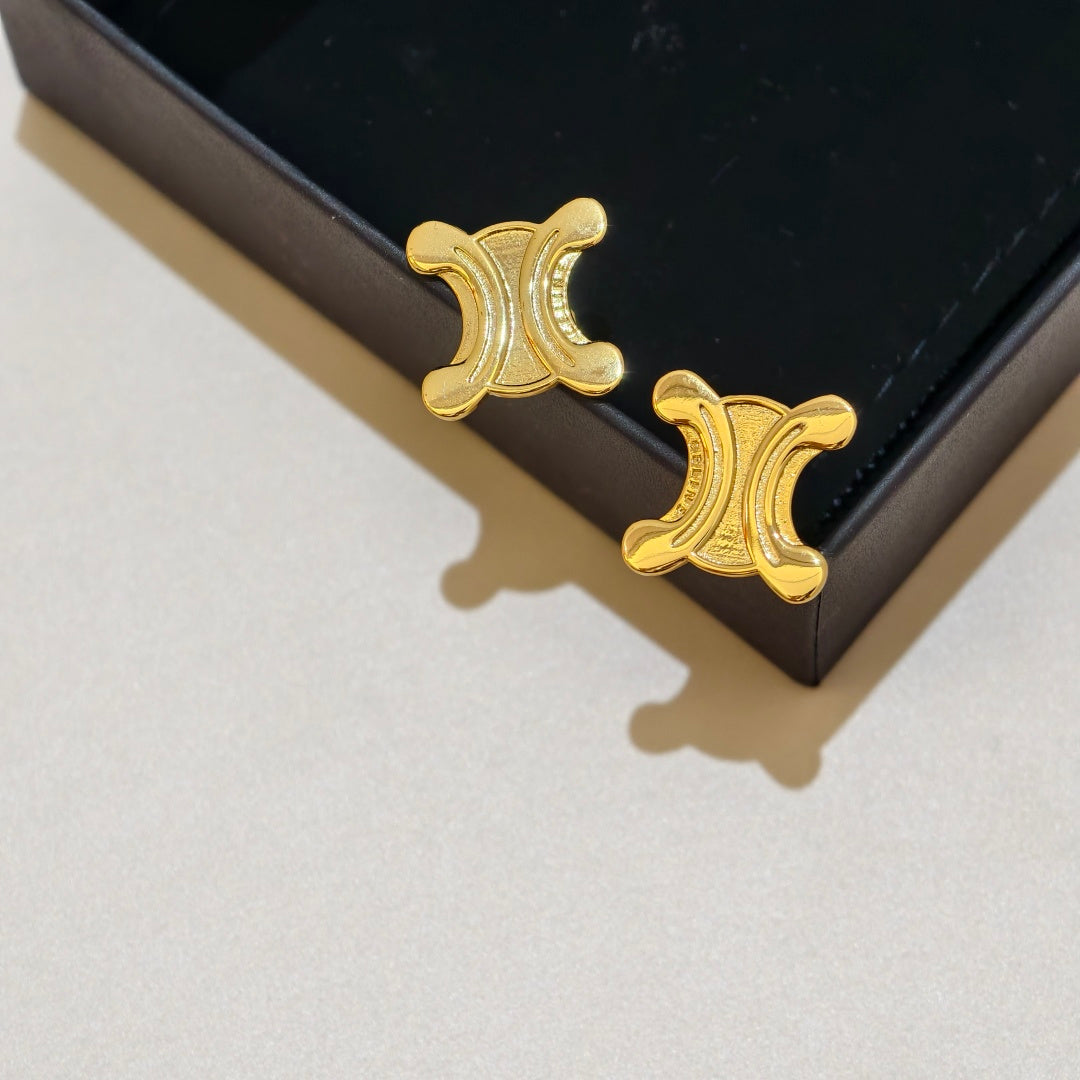 A pair of gold-tone stud earrings with a crescent moon and abstract curved motif, resting on a black jewelry box against a light beige background.