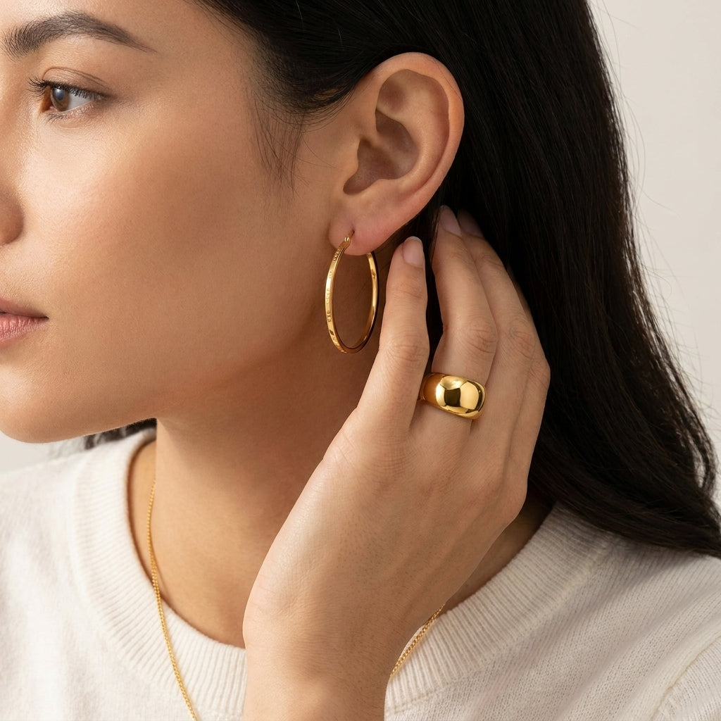 A close-up lifestyle shot of a woman wearing a polished gold hoop earring and a matching gold ring, shown against a soft beige background.