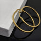 A pair of gold hoop earrings with engraved text, resting on a white textured box against a glossy black reflective surface.