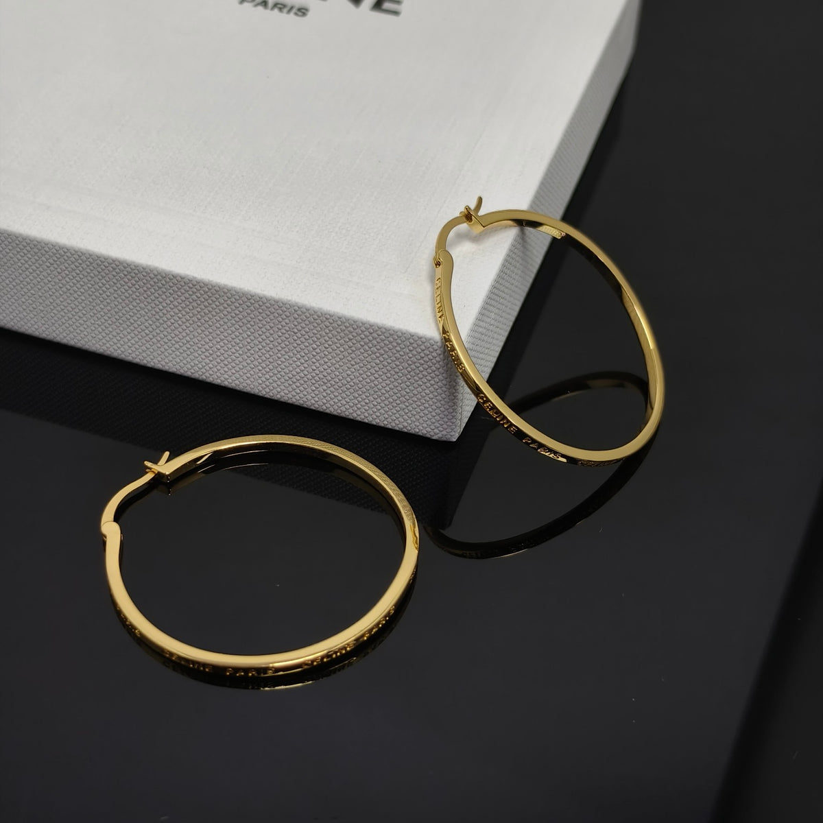 A pair of gold hoop earrings with engraved text, displayed on a glossy black surface next to a white textured box.