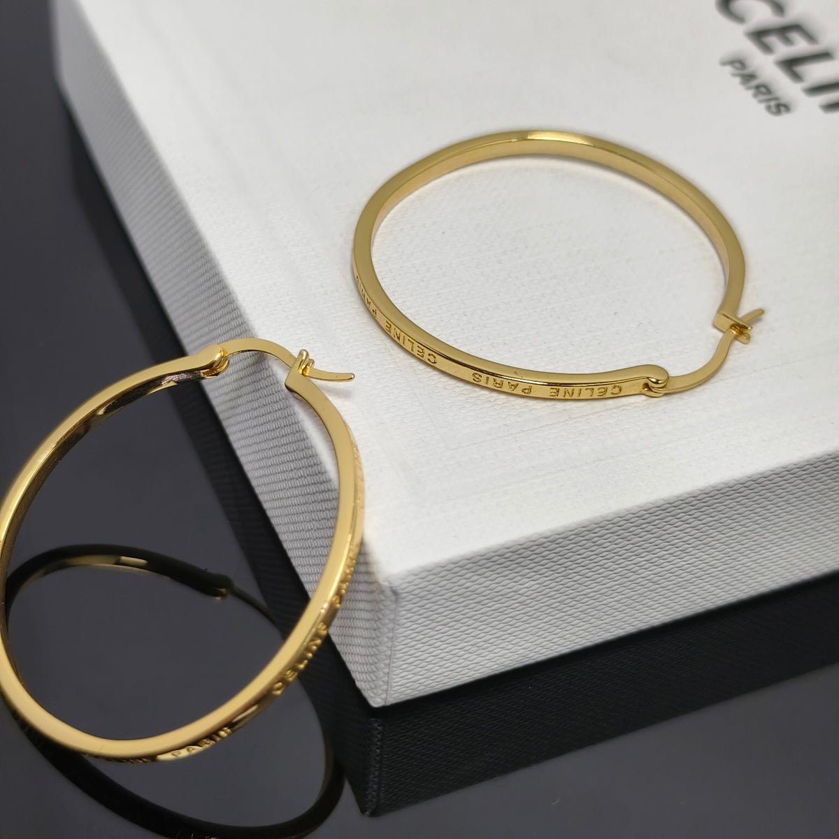 Gold hoop earrings displayed on a white textured box with the brand name CELINE PARIS partially visible, alongside a matching gold bangle bracelet.