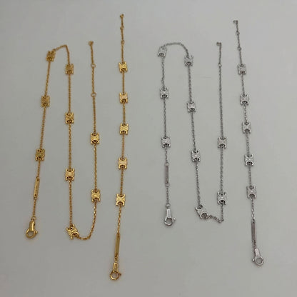 Two delicate chain necklaces laid flat on a light gray background, one in yellow gold and one in silver, both featuring repeating decorative links and lobster clasp ends.
