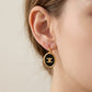 A close-up of a model's ear wearing a gold hoop earring with a black oval charm featuring a gold interlocking CC logo, indicating the Chanel brand.