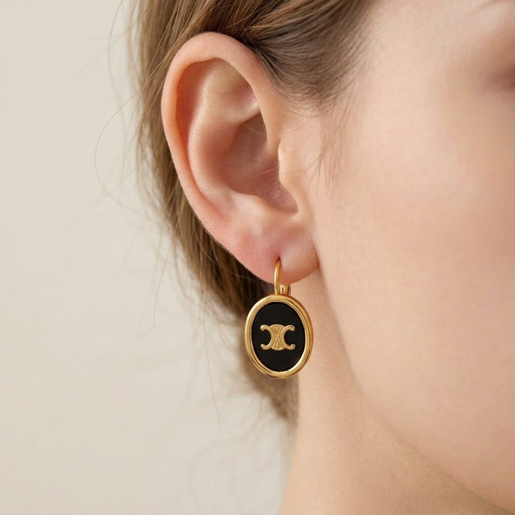 A close-up of a model's ear wearing a gold hoop earring with a black oval charm featuring a gold interlocking CC logo, indicating the Chanel brand.