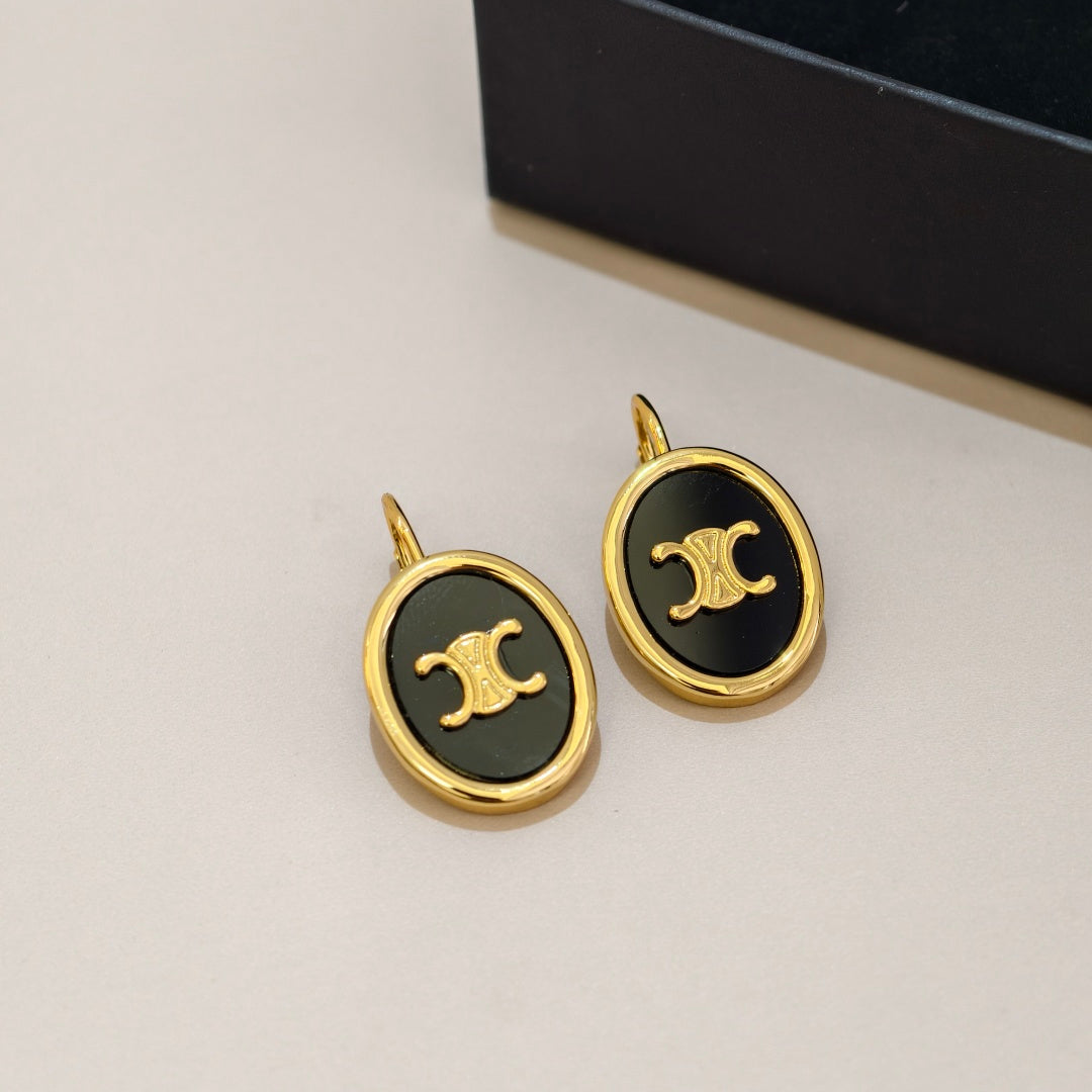 A pair of oval stud earrings with gold-tone metal frames and black enamel centers featuring a gold interlocking double-C logo, shown on a light beige surface with a dark box in the background.