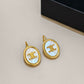 A pair of gold oval drop earrings with iridescent white mother-of-pearl inlays featuring a gold triple-C logo, displayed on a neutral beige surface with a black box in the background.