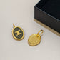 Two oval gold-tone earrings with black enamel inserts and a gold logo emblem, displayed next to a black presentation box on a light beige surface.