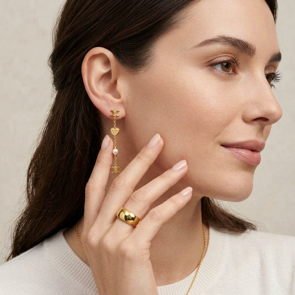 A close-up of a woman wearing a delicate gold dangle earring with a small gold heart, a pearl, and a gold bird, paired with a gold ring and necklace.