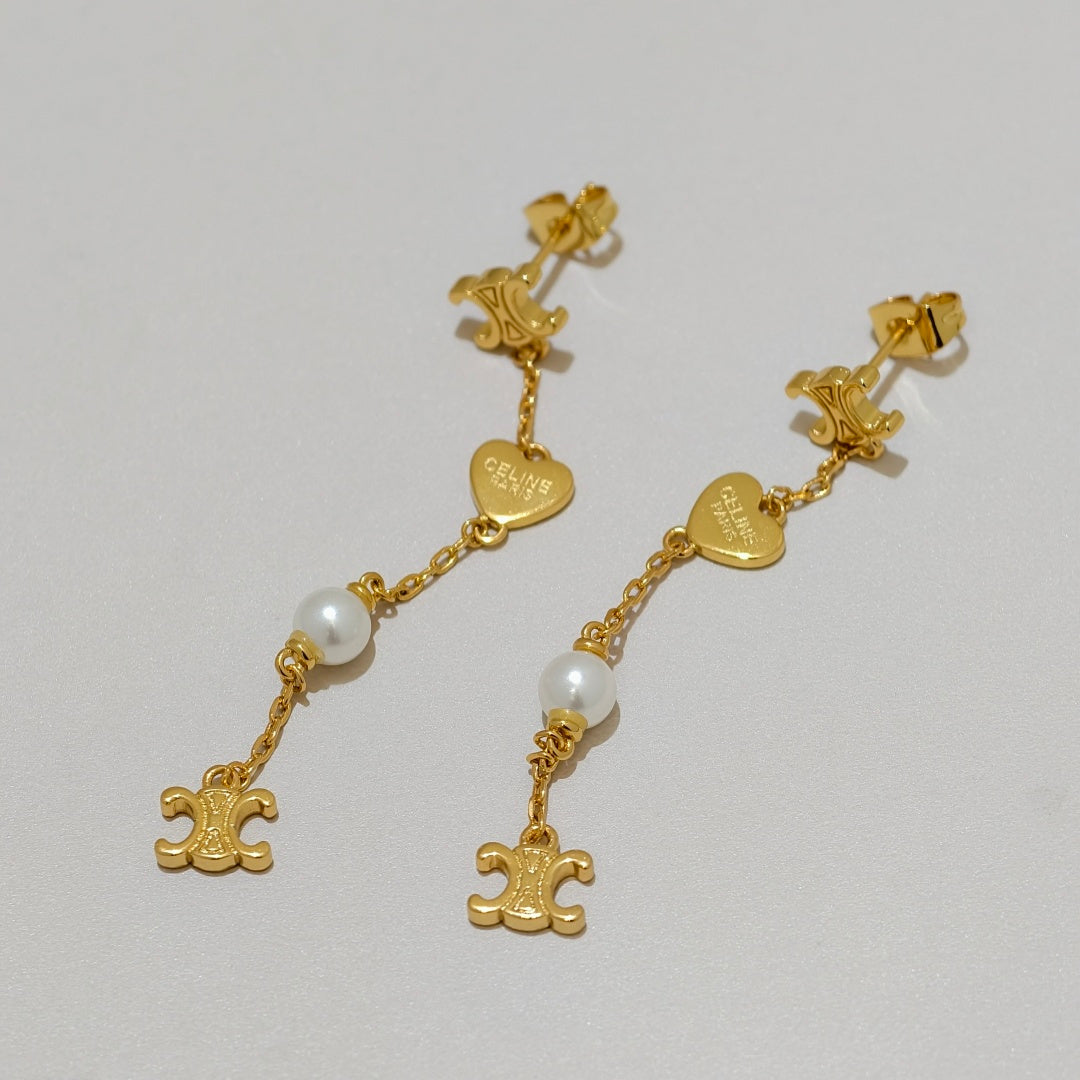 A pair of gold dangle earrings with heart-shaped charms, white pearl beads, and fleur-de-lis charms, displayed on a light gray background.