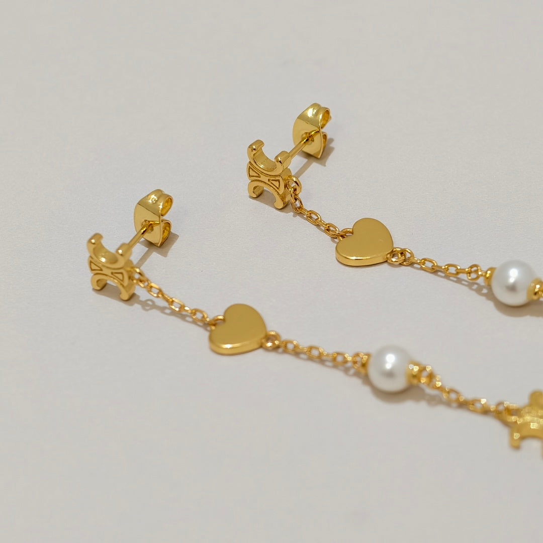 Gold dangle earrings with heart charms, star details, and white pearl beads on a light gray background.