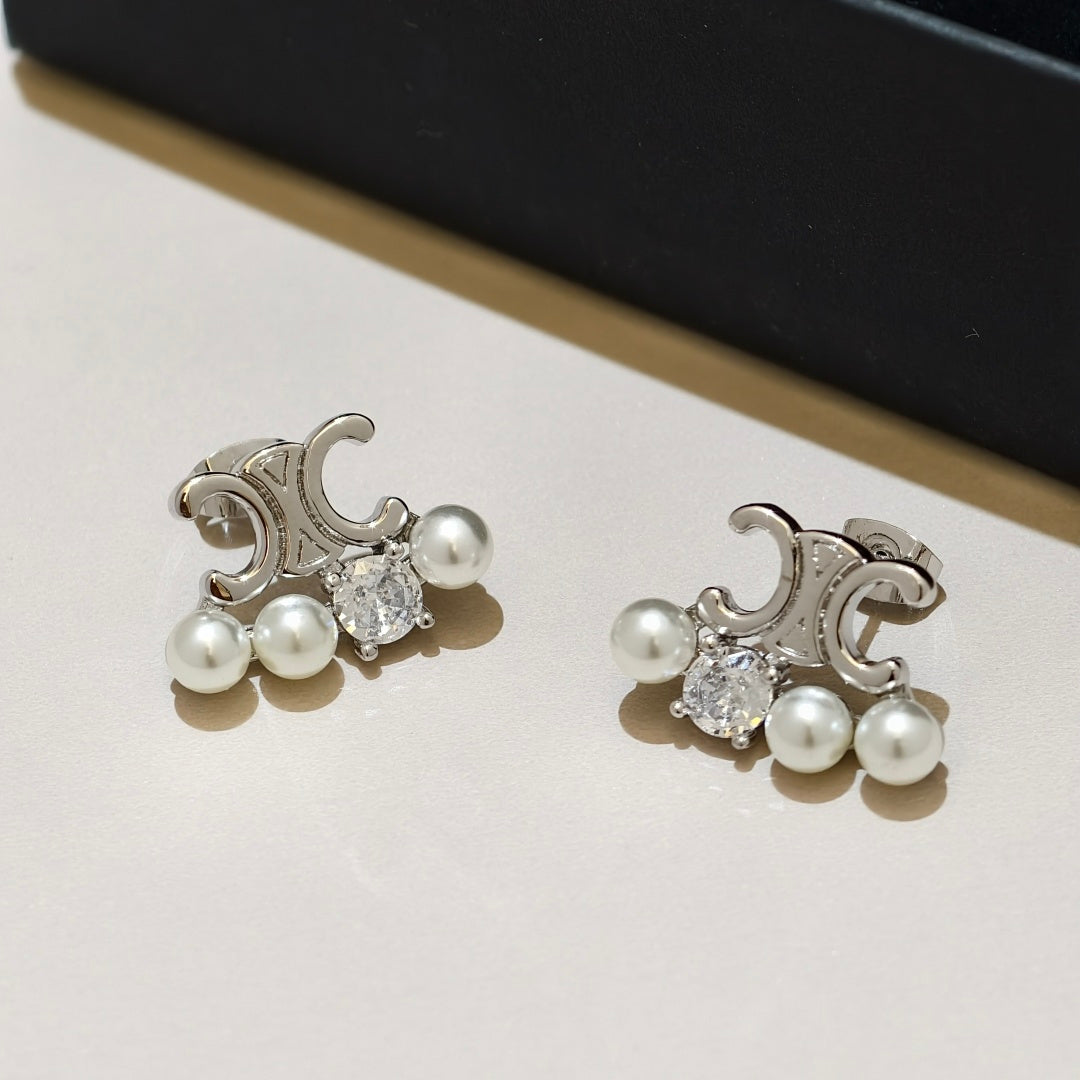 A pair of silver-toned stud earrings with three white pearls and a clear round-cut gemstone, displayed on a light beige surface with a dark box in the background.