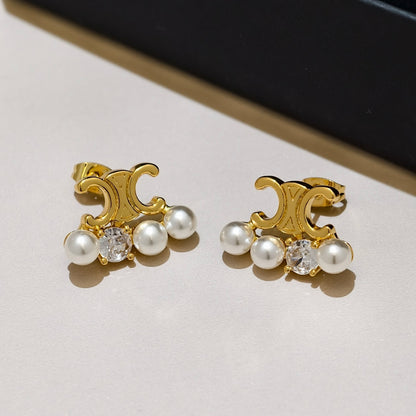 A pair of gold-toned clip-on earrings with a curved ornate motif, three white faux pearls, and a clear round crystal in the center, photographed on a light beige surface with a dark navy background.